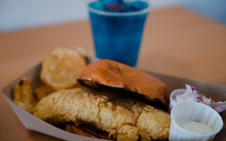 Buoy Shack's fried fish sandwich is shown with a toasted bun, tartar sauce, housemade chips, and cole slaw.