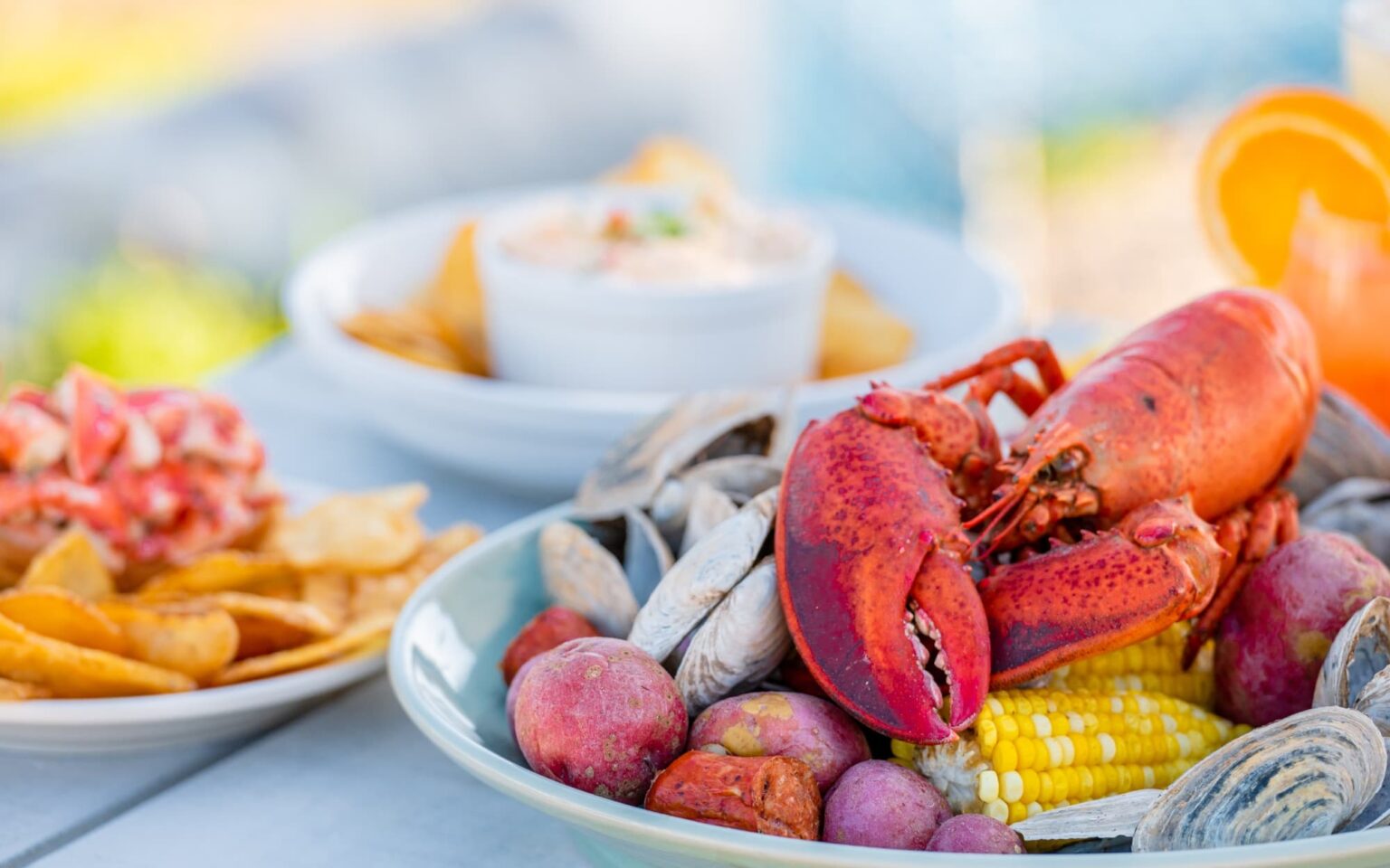 A whole lobster sits on top of corn, red potatoes, and whole clams. In the background, lobster dip is shown along with a lobster roll and housemade chips.